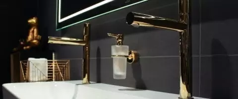 Goldene Badarmaturen an einem Waschbecken und in einem Bad mit schwarze Wandfliesen.