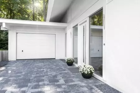Garage mit weißem Garagentor an einem Einfamilienhaus mit einer Einfahrt aus Pflastersteinen.