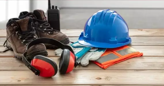 Auf einer Holzarbeitsplatte stehen Arbeitsschuhe und Arbeitsschutzkleidung wie ein blauer Arbeitshelm, Handschuhe und ein Gehörschutz.