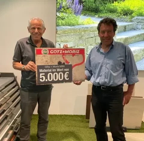 Zwei glückliche Männer halten einen 5000€ Check von Götz + Moriz in der Hand.