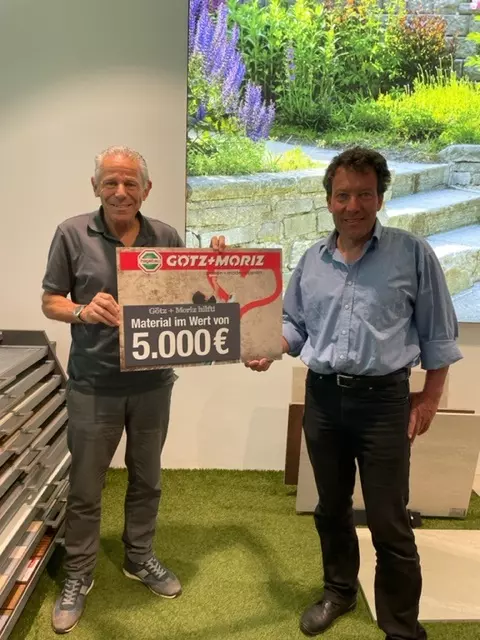 Zwei glückliche Männer halten einen 5000€ Check von Götz + Moriz in der Hand.